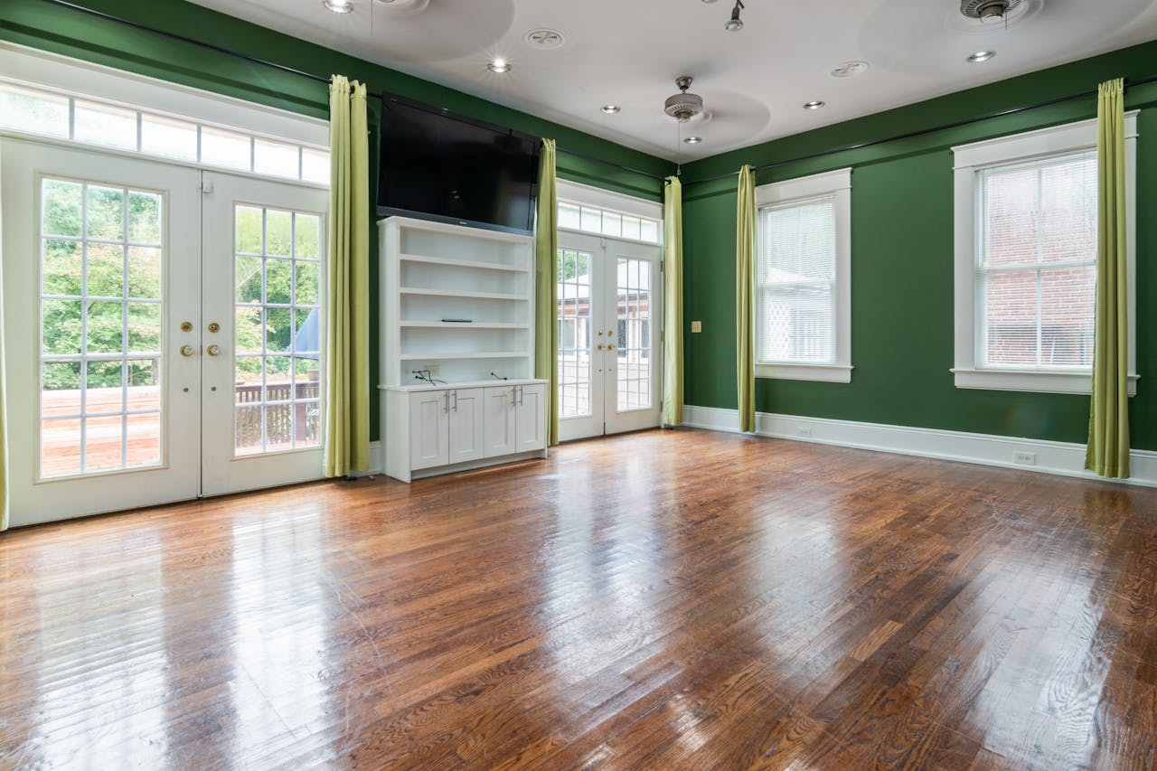 Empty sunlit room with hardwood floor, large windows, and green walls.